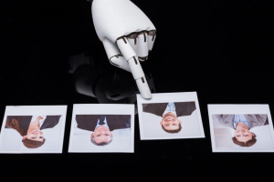 Image of white robotic hand pointing at a polaroid of a man in a suit, with two other polaroids to the left and one to the right. The robot is selecting the individual in the picture they are pointing at. 